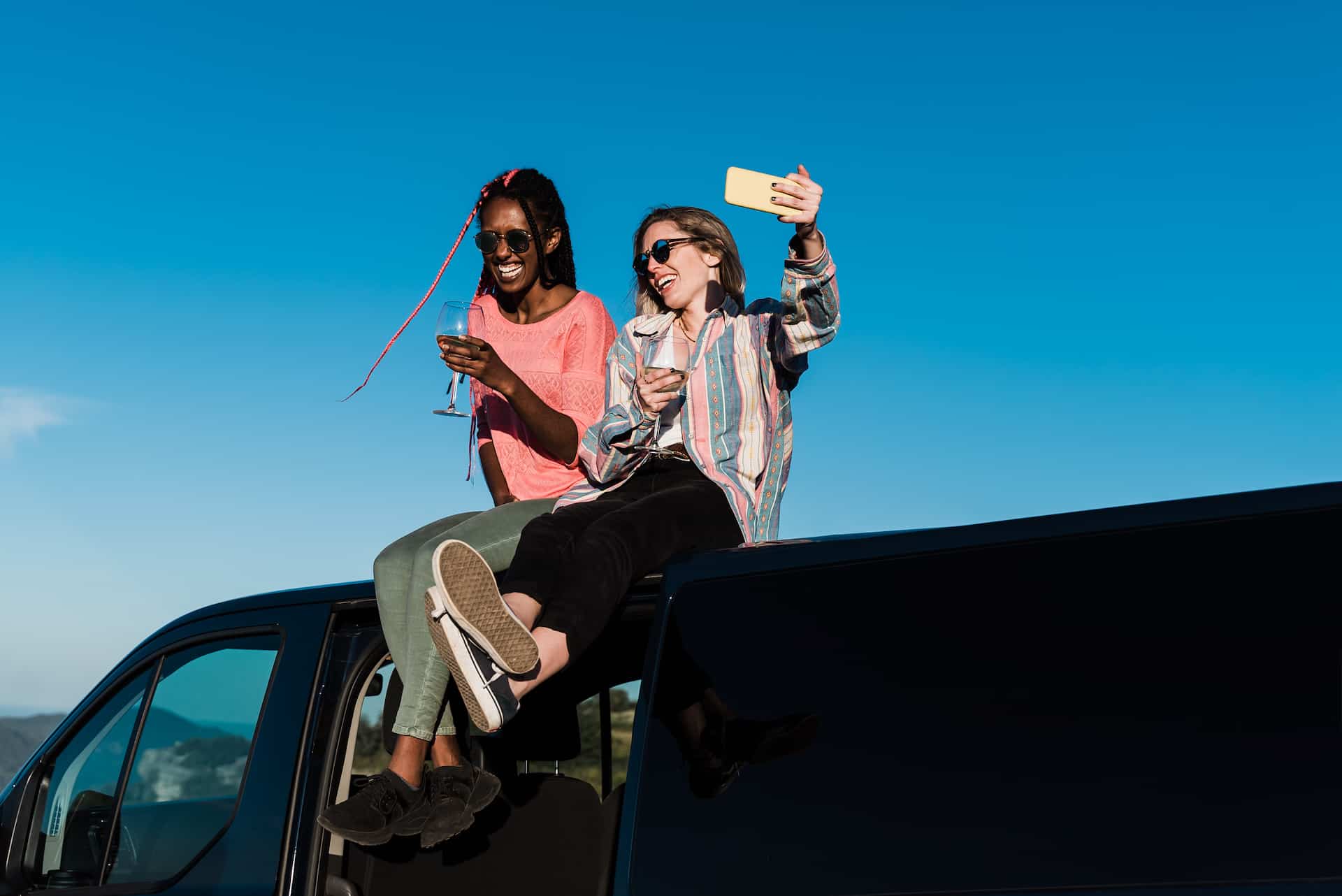Happy multiracial people sitting on top of camper van roof at summer sunset - Travel, lifestyle and freedom concept
