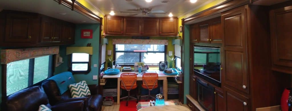 The inside of an RV is shown. There are dark wood cabinets, a desk with red desk chairs and two leather chairs with pillows on them.