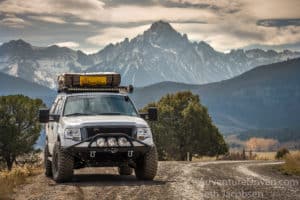An overlanding truck with a roof rack and flood lights is pictured in front of a mountain range