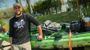 Adam James is wearing a Battle Born Batteries long sleeve shirt, and standing and smiling in front of a green kayak.