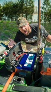 Adam James is wearing a Battle Born Batteries long sleeve shirt standing with his mouth open over a 100Ah Battle Born Battery in his kayak.