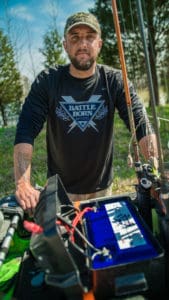 Adam James is wearing a Battle Born Batteries long sleeve shirt and standing over a 100Ah Battle Born Battery in his kayak.