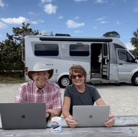 Two people on laptops with an RV in the background behind them