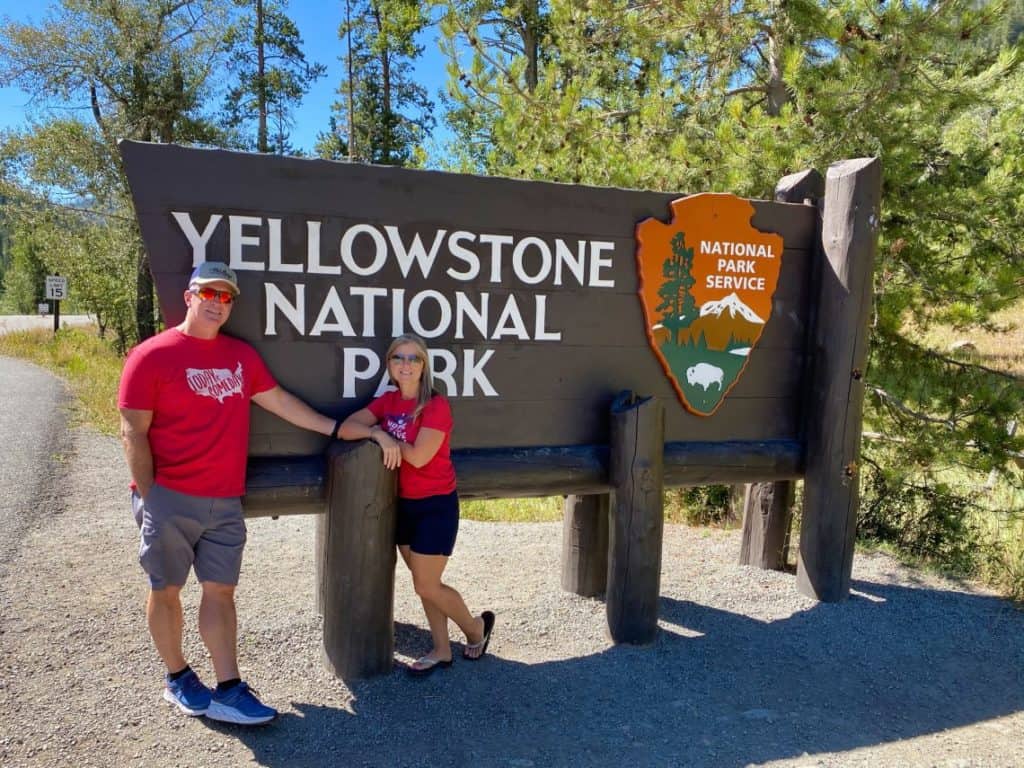 A couple stands in front of the Yellowstone National Park sign. 