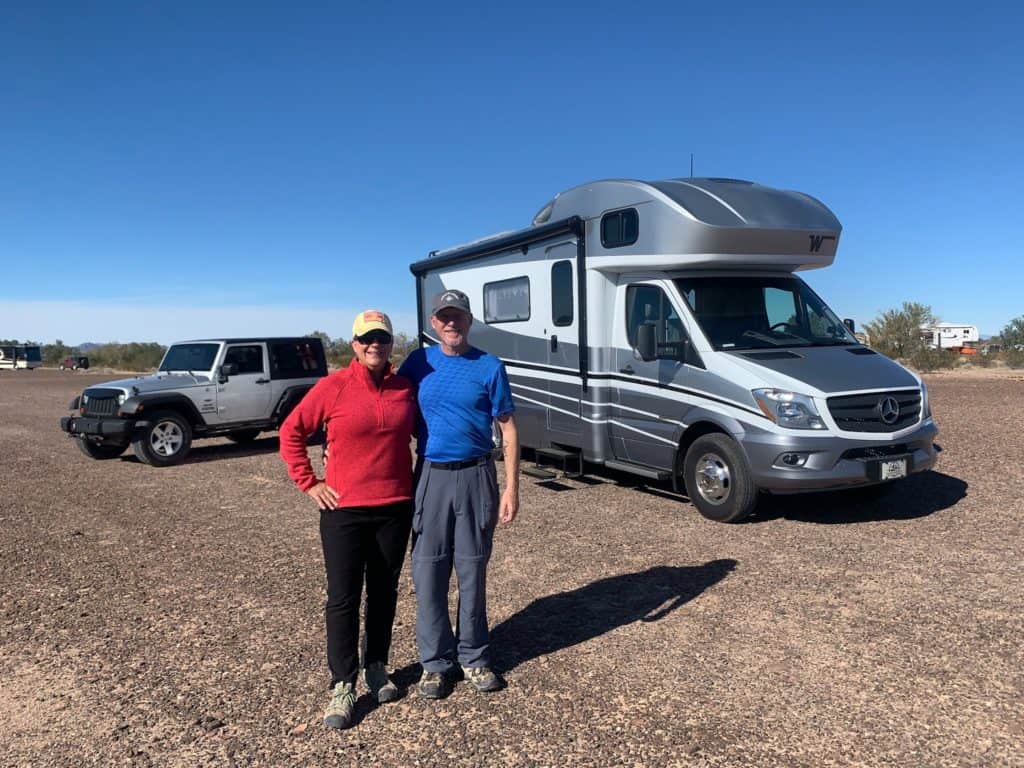 Two people standing in front of a small motorhome and jeep