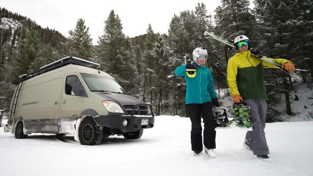 Two people carrying their snow skis outside of a van in the snow.
