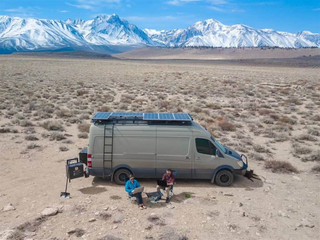 Overhead shot of a Sprinter van with solar panels on top parked in the middle of the desert. There are two people working on laptops in front of the van.