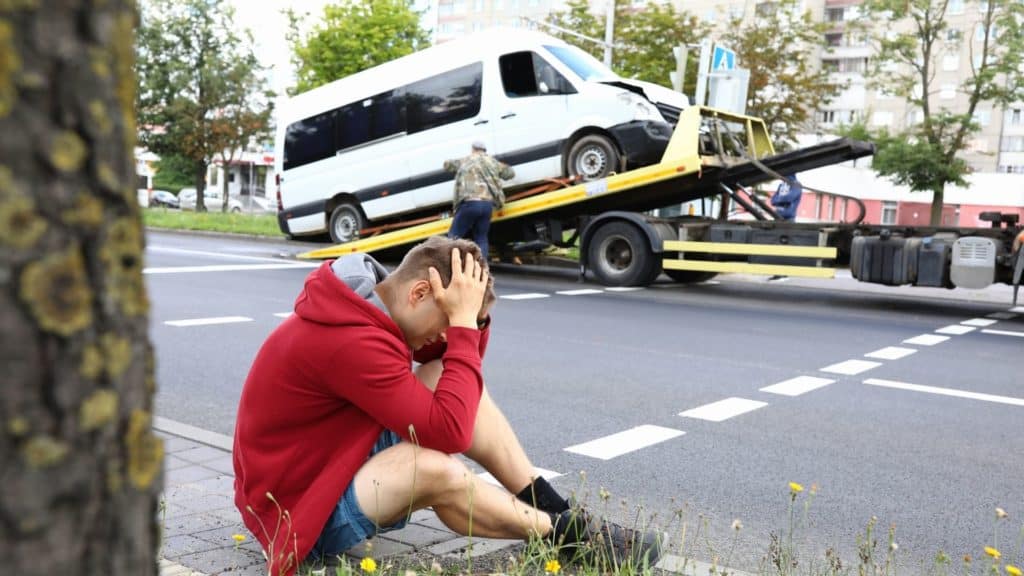 man holding head after van accident, van is being towed in the background