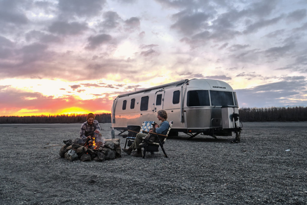 Two people sitting around a campfire with an Airstream trailer in the background