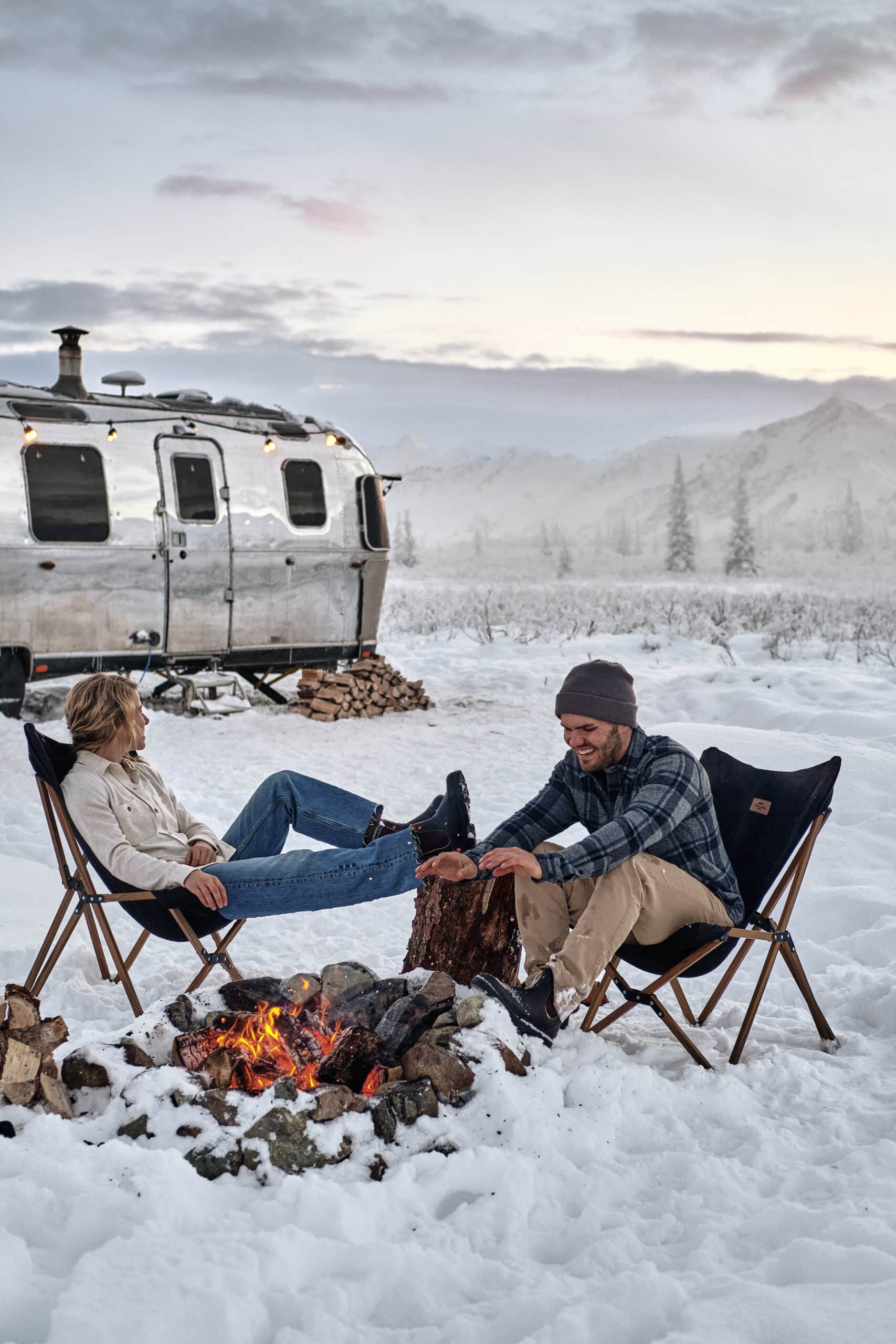Two People with a Campfire in the Snow in Front of Their Airstream Camper in Alaska