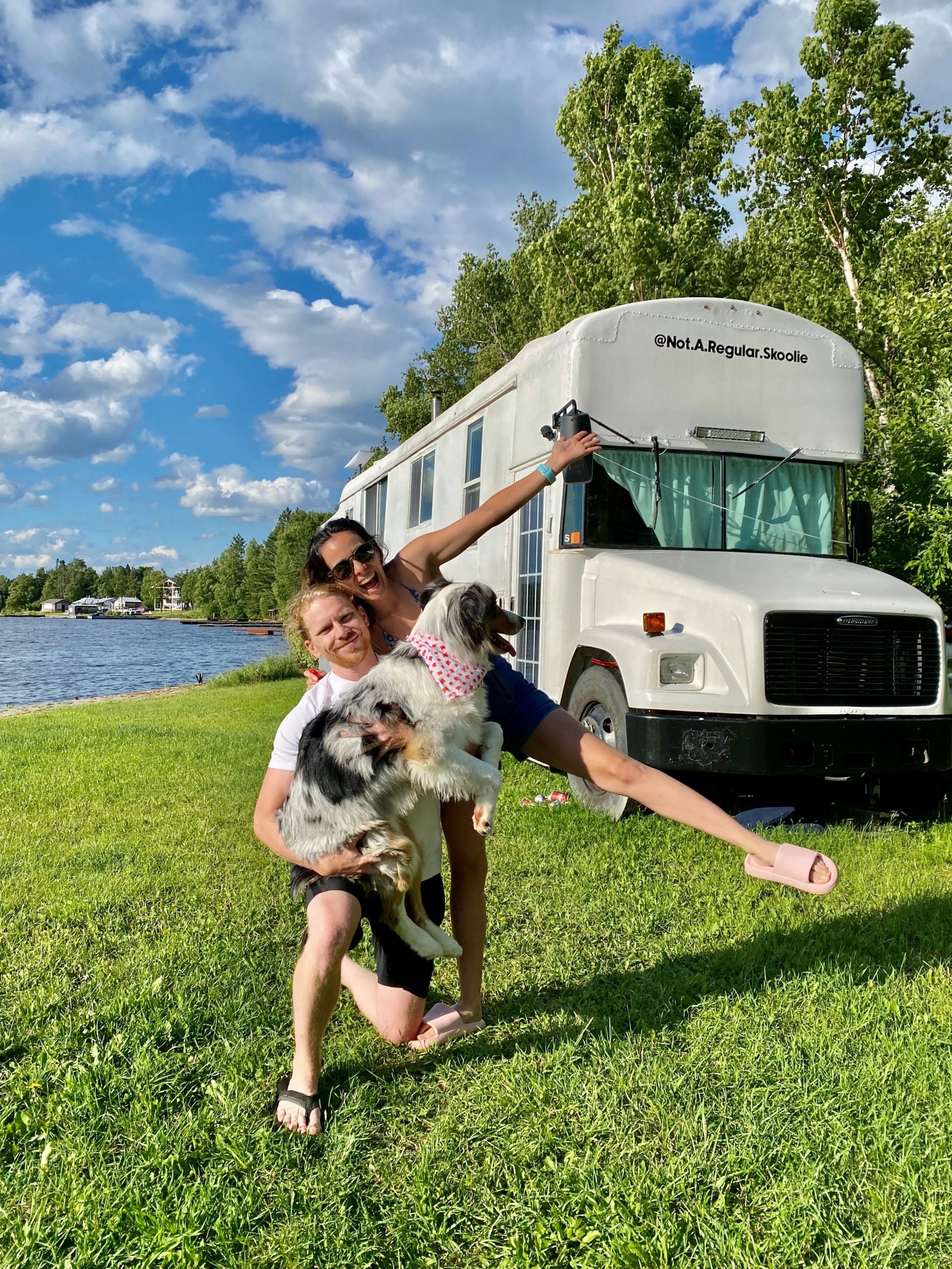 Gabi and Tyler from Not a Regular Skoolie standing with their dog outside of their bus in grass next to a lake