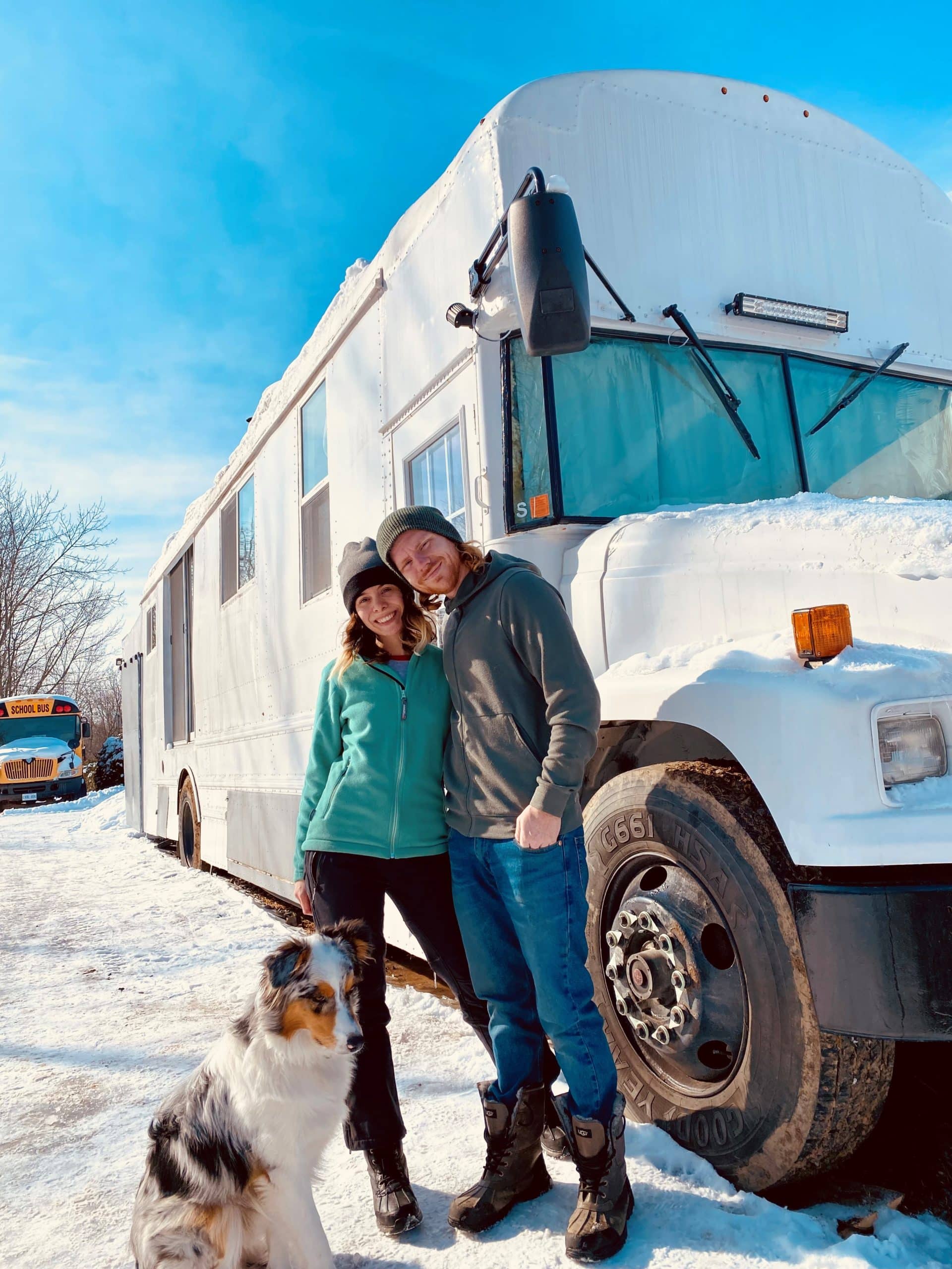 Gabi and Tyler from Not a Regular Skoolie standing with their dog outside of their bus in the snow