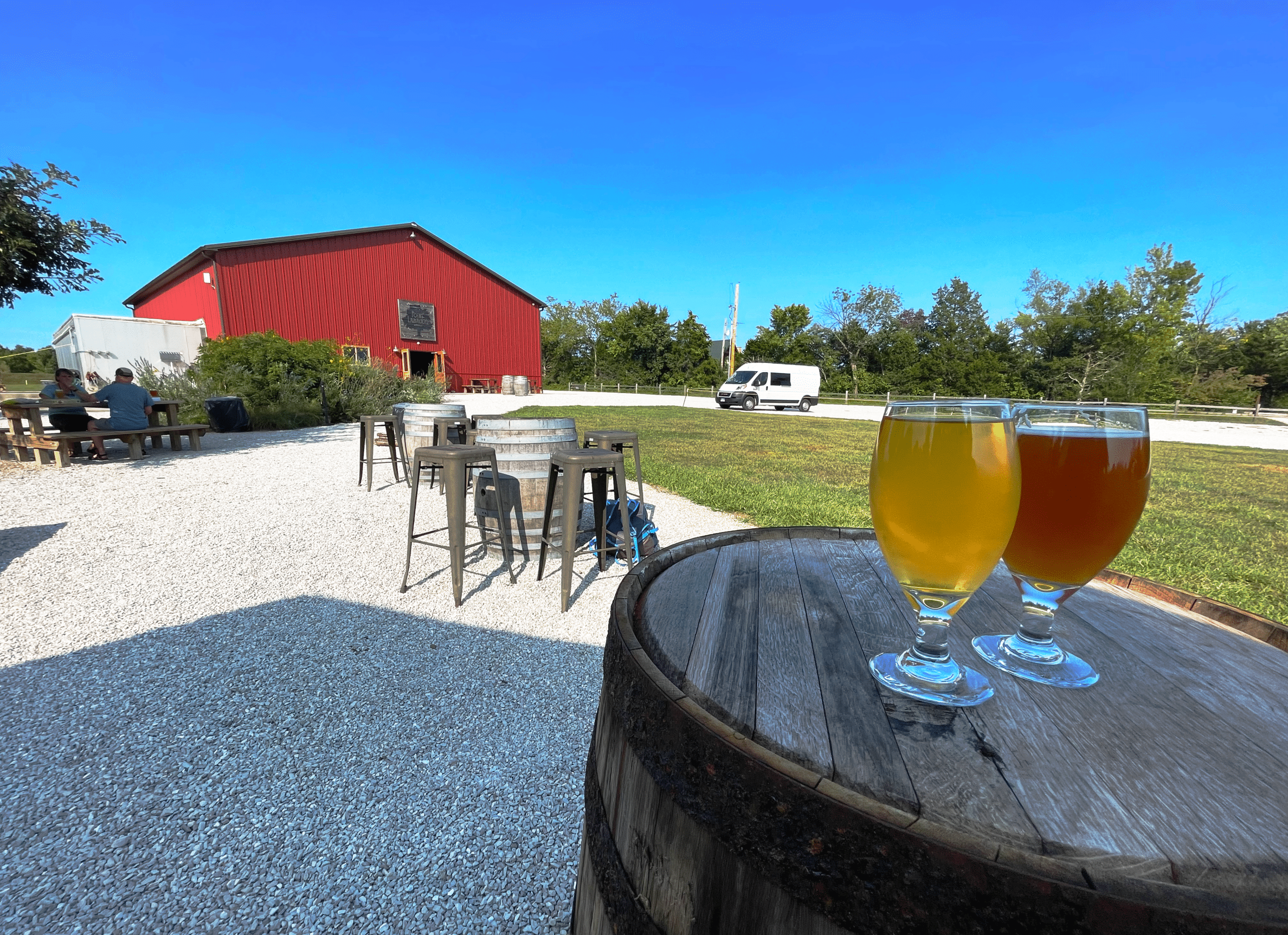 Two beer glasses on top of a wine barrel with a barn in the background