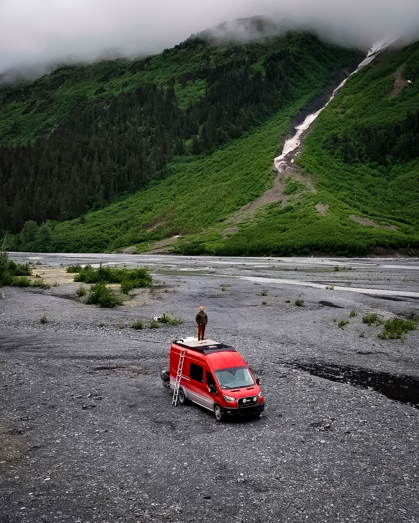 A drone shot of a red van boondocking next to a river and mountains