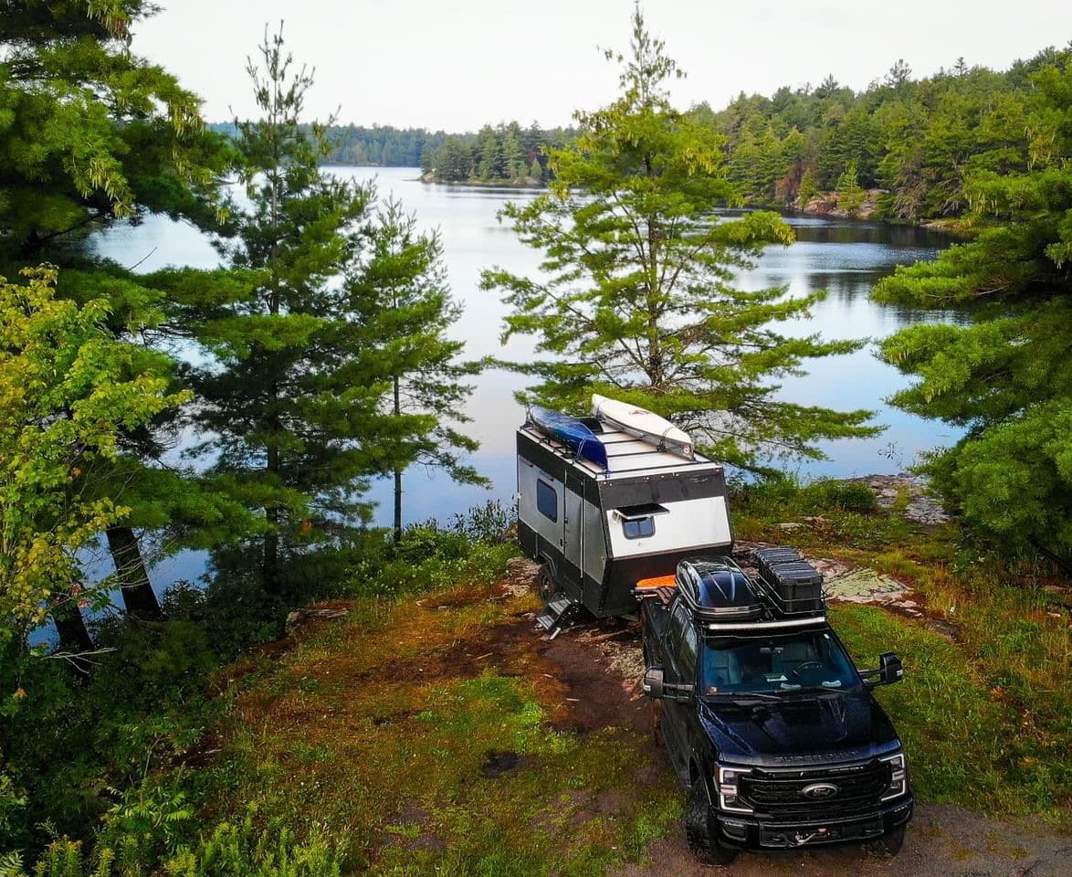 A truck towing an RV with canoes on top is parked next to a body of water