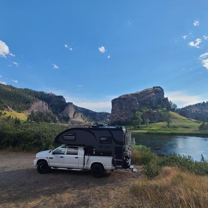 A white truck has a cirus 820 truck camper on the back while parked next to a body of water