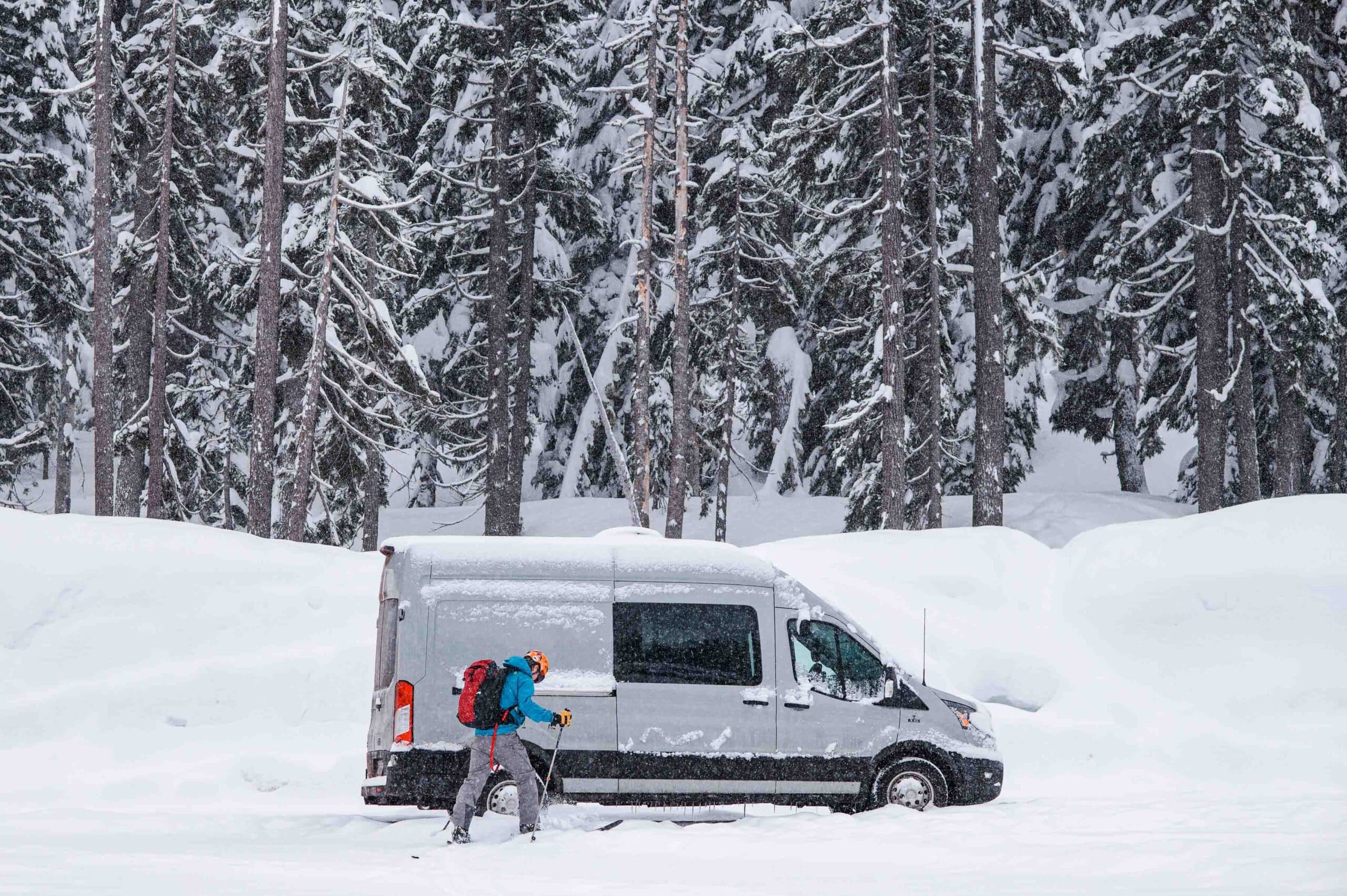 A man wearing snow skis outside of a van boondocking while covered in snow